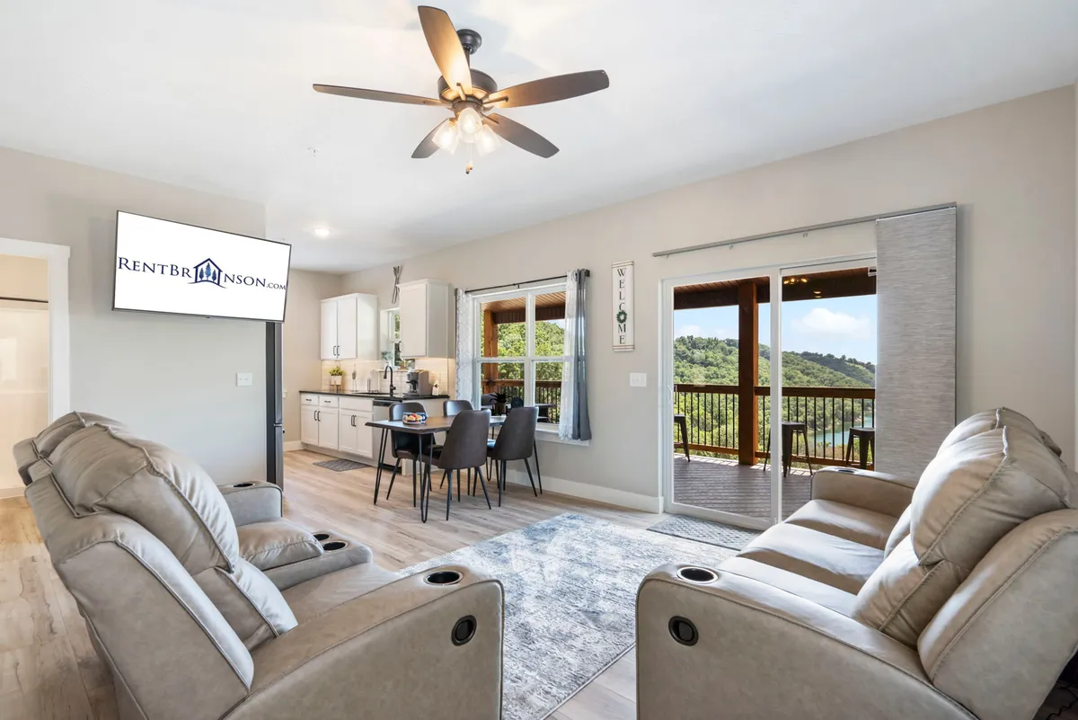 A bright, inviting interior with reclining chairs, a dining table set for shared meals, large windows allowing in natural light and scenic views, and the kitchen area visible in the background. The space feels ready for both relaxation and lively conversation.