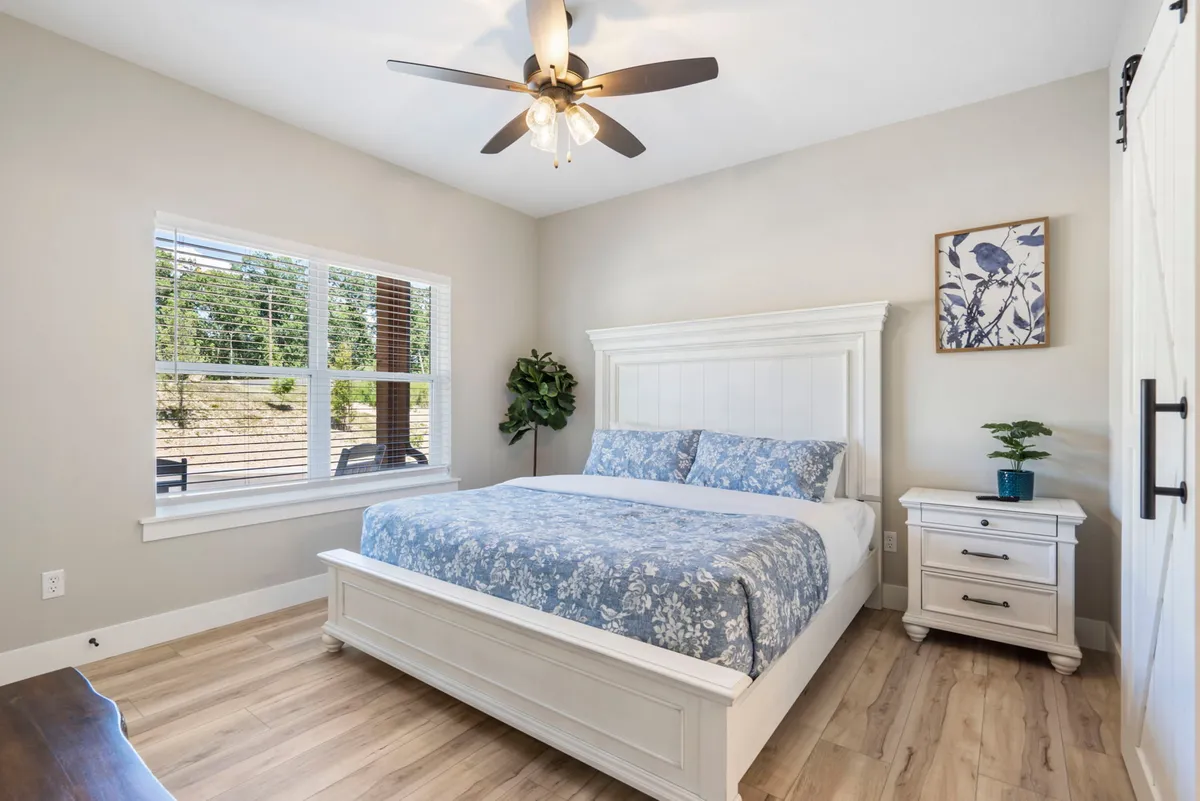 A restful bedroom with floral bedding, a nightstand topped by a leafy plant, and a window letting in soft morning light. The overall mood is calm and cozy, perfect for quiet moments or reading by the window.