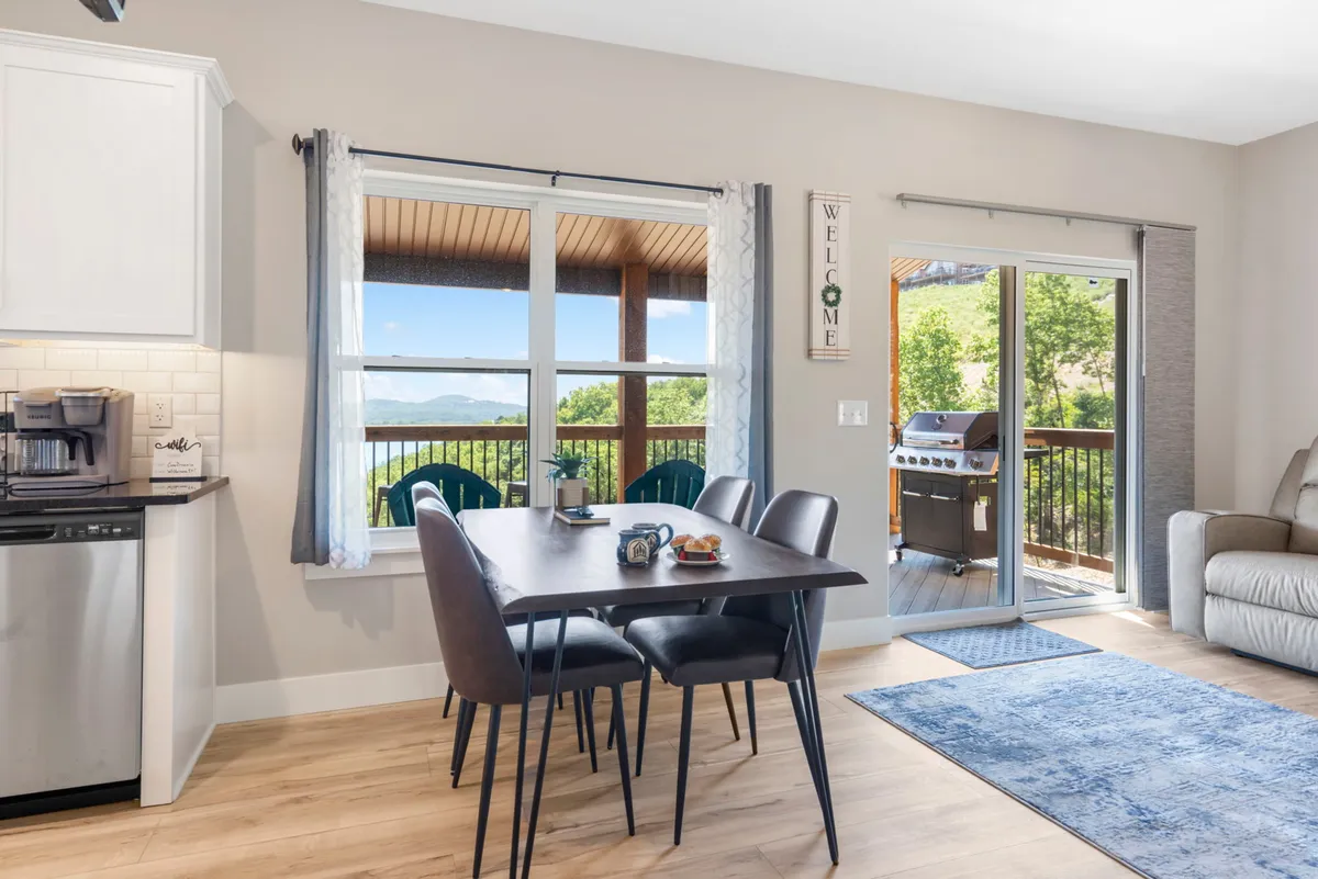 The image presents a dining table adjacent to large windows leading out to an outdoor deck, where chairs and a grill are visible. It highlights the transition from indoor dining to outdoor entertaining, echoing the invitation for lakeside feasts and gatherings.