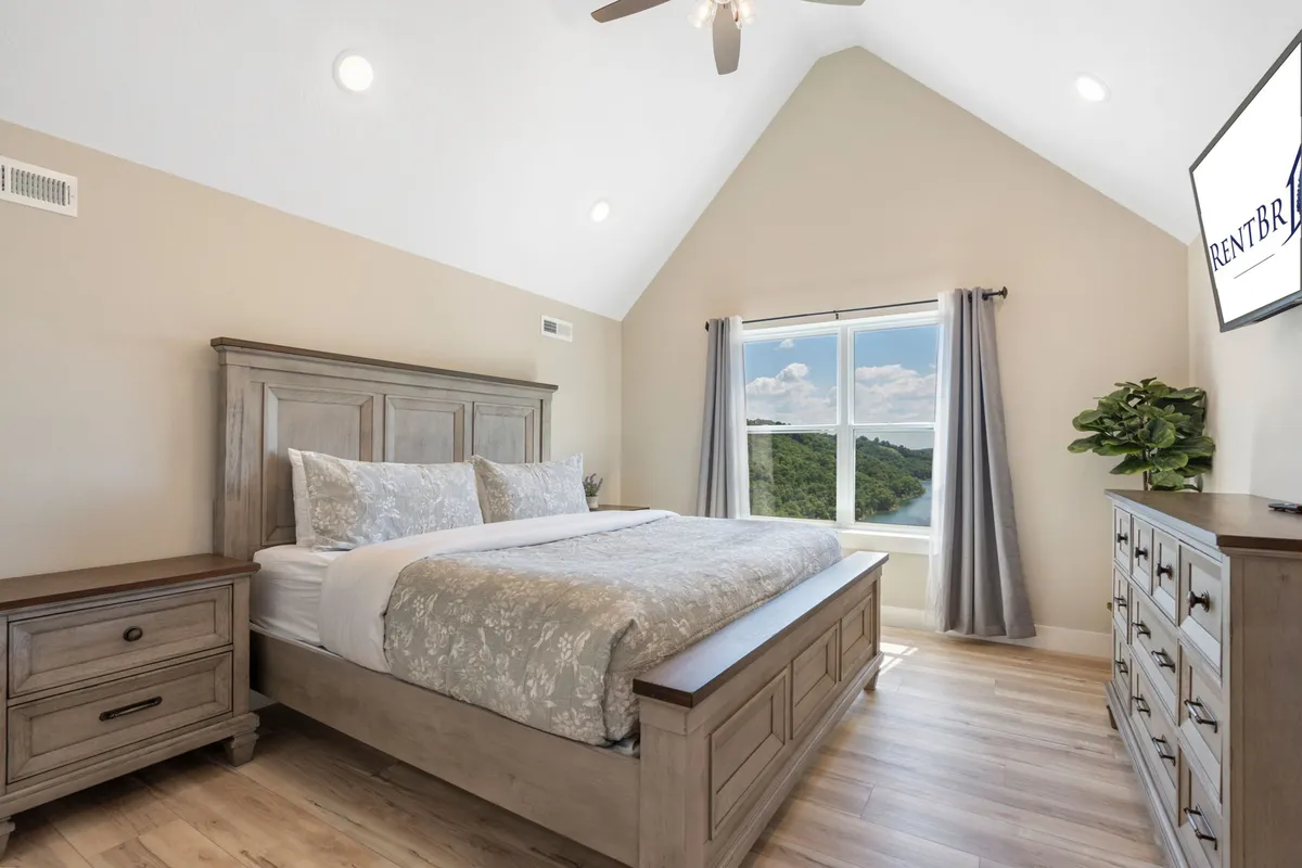 A serene bedroom featuring a king-sized bed, a large window offering natural light and partial views, and a wooden dresser. The room exudes a calm, restful atmosphere ideal for relaxation after a day of adventure.