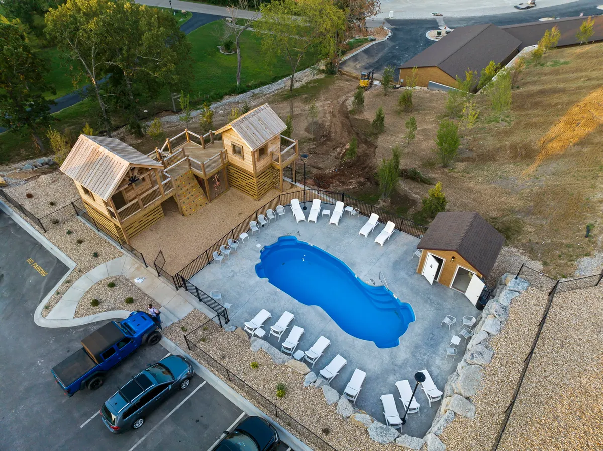 A bright, inviting outdoor pool area with a large swimming pool, lounge chairs, and a playground structure adjacent to the pool. Children can be seen playing while families relax and sunbathe nearby, creating a vibrant, family-friendly atmosphere.