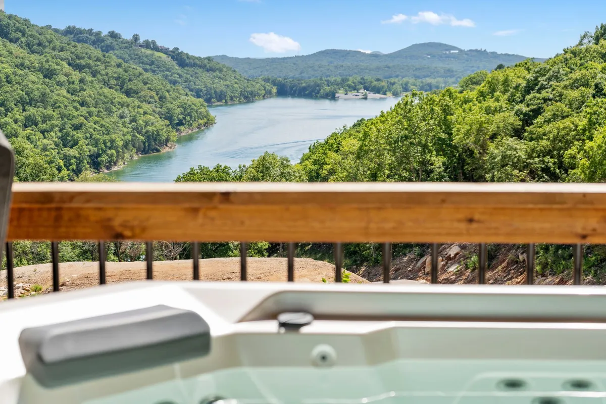 A serene hot tub on a wooden deck overlooking a picturesque lake and rolling hills. The expansive view highlights both the hot tub area and the tranquil outdoor setting, perfect for unwinding at any time of day.
