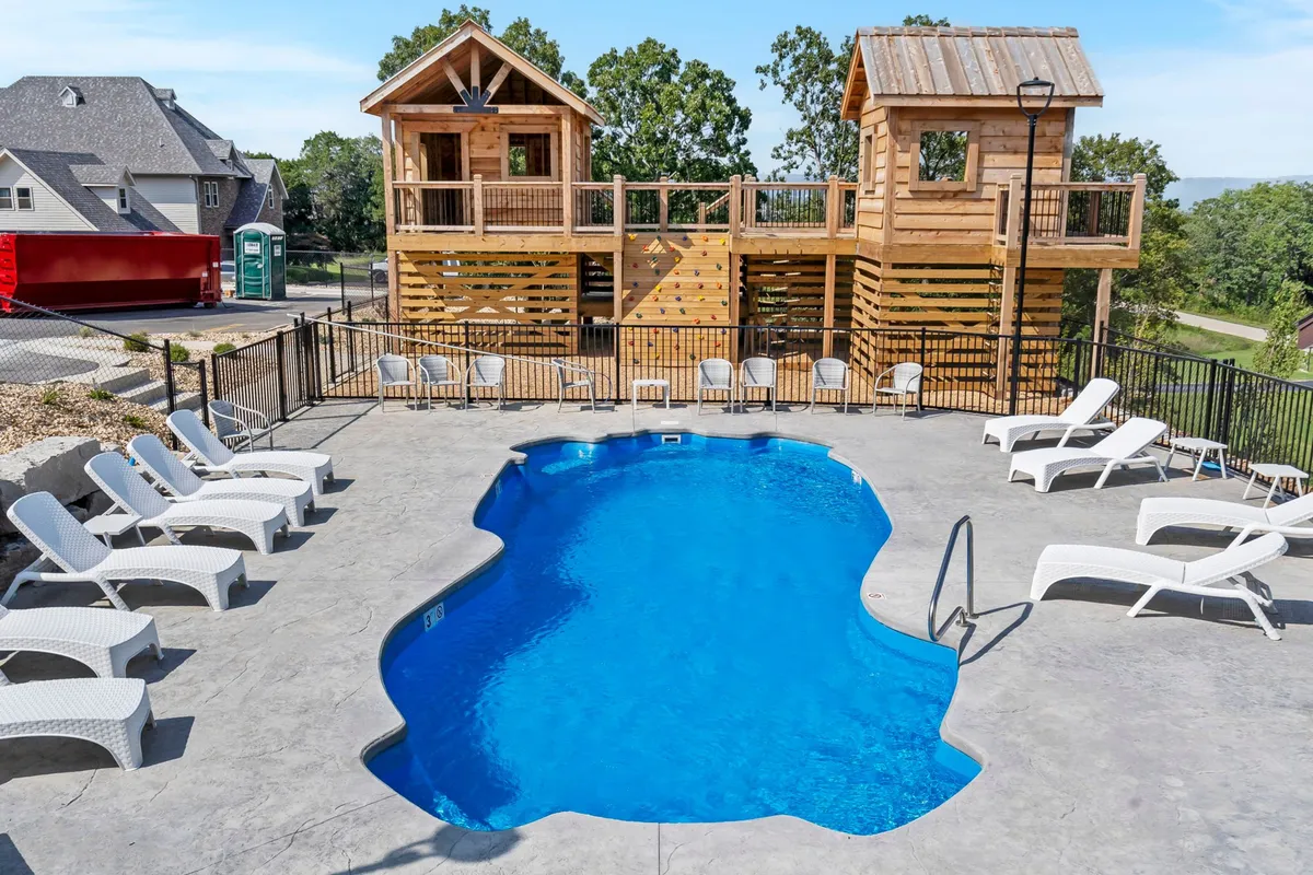 This lively exterior scene highlights a sparkling swimming pool surrounded by wooden playhouses and comfortable lounge chairs. Children can be seen playing and having fun, while families gather poolside, enjoying the sunshine and relaxed atmosphere. The backdrop of resort buildings and landscaping adds to the cheerful, family-friendly mood.