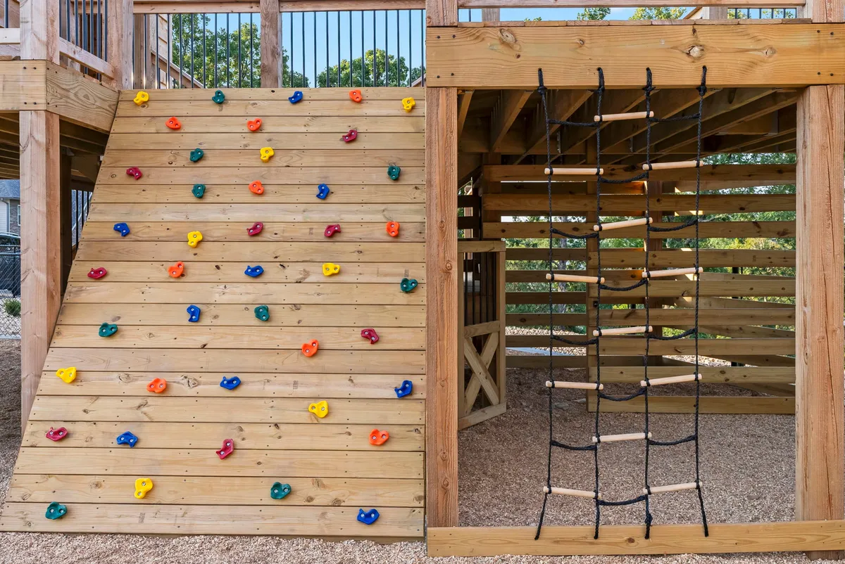 This energetic exterior image features a colorful climbing wall and rope ladder as part of an outdoor playground. Children are actively climbing and playing, surrounded by greenery. The joyful expressions and playful activity evoke a sense of adventure and outdoor exploration for guests of all ages.