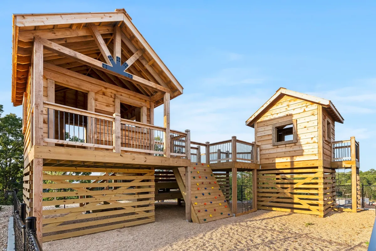 A whimsical exterior playground scene featuring a treehouse structure, climbing wall, slide, and lush greenery. Children are actively playing and exploring, bringing energy and adventure to the communal outdoor space.